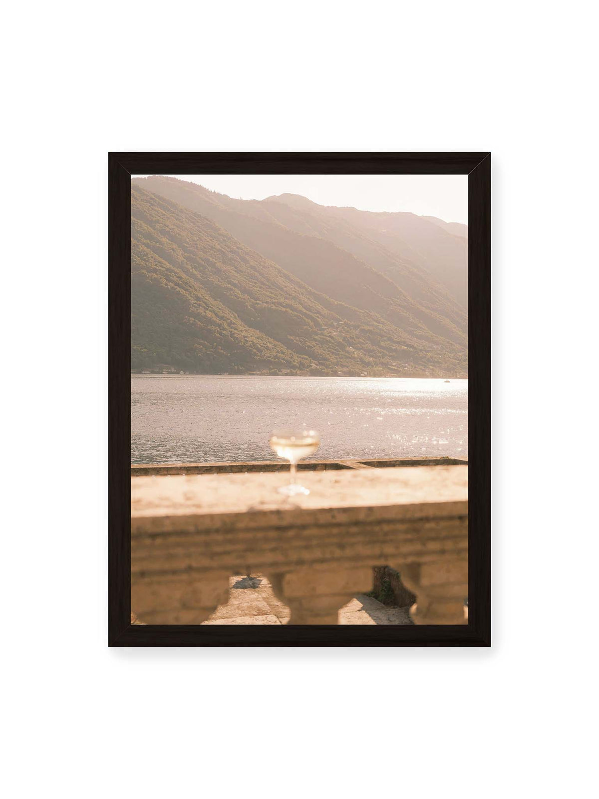 30x40 cm, schwarzer Rahmen – Einzelnes Glas auf heller Steinfläche vor glitzerndem Wasser und bewaldeten Berghängen.