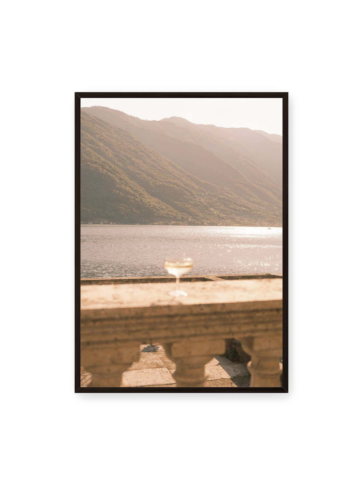 70x100 cm, schwarzer Rahmen – Einzelnes Glas auf heller Steinfläche vor glitzerndem Wasser und bewaldeten Berghängen.