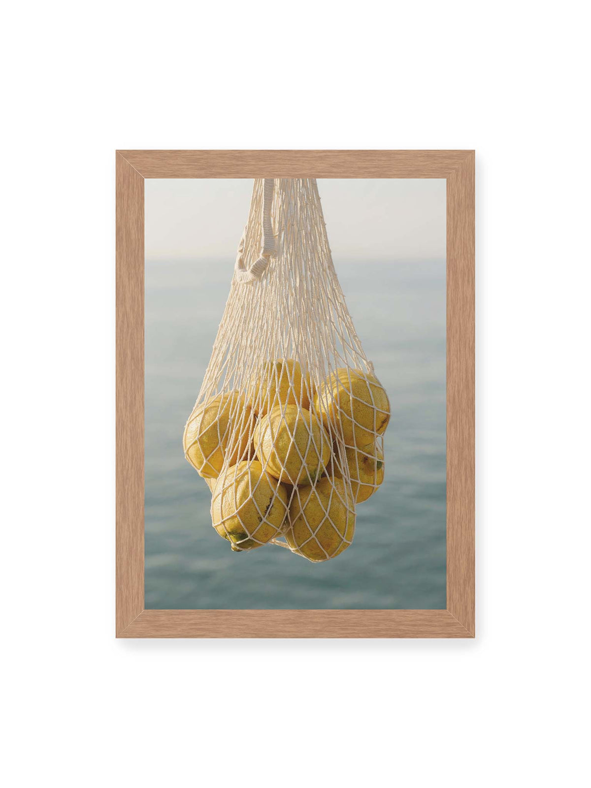 21x30 cm, Eichenrahmen – Gelbe Zitronen in einem hellen geknüpften Netz vor einer weich verschwimmenden blauen Wasserfläche.