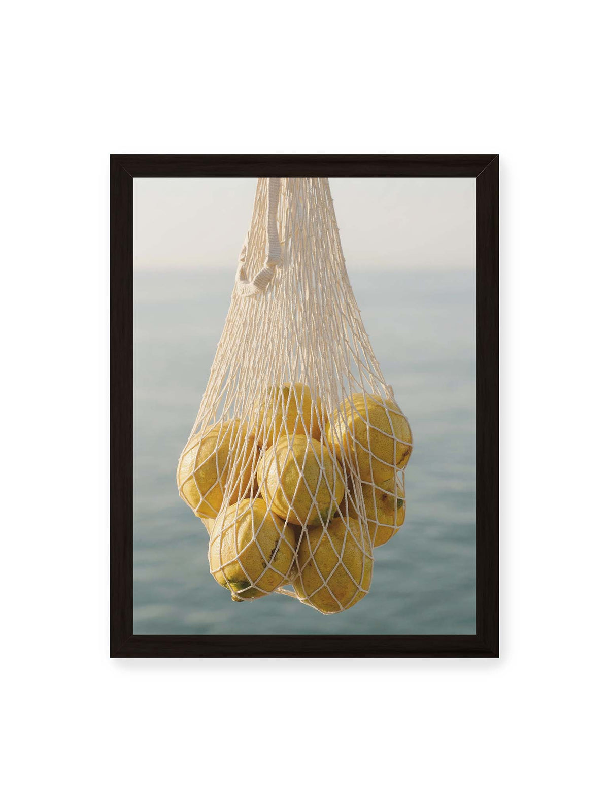 30x40 cm, schwarzer Rahmen – Gelbe Zitronen in einem hellen geknüpften Netz vor einer weich verschwimmenden blauen Wasserfläche.