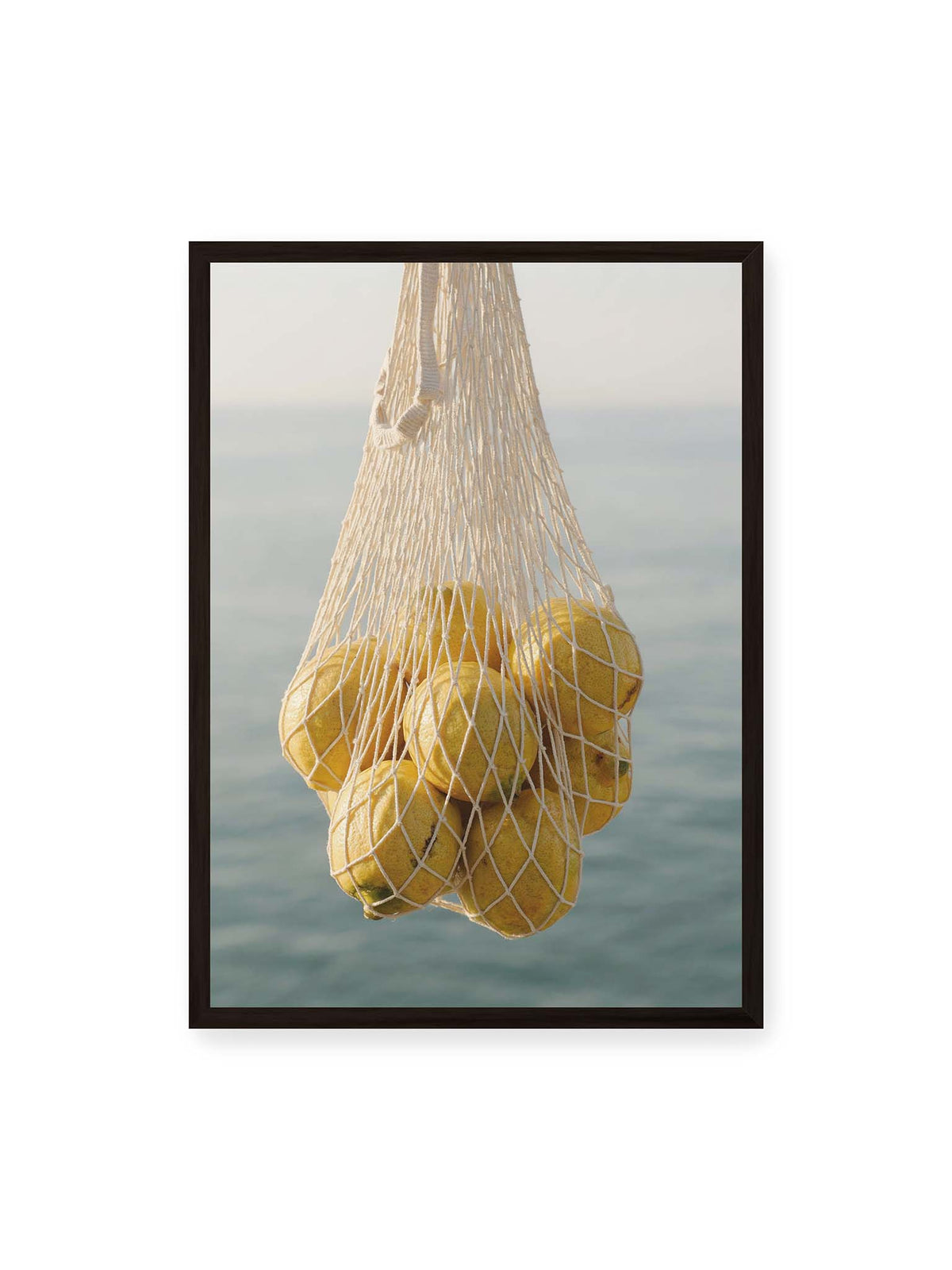 50x70 cm, schwarzer Rahmen – Gelbe Zitronen in einem hellen geknüpften Netz vor einer weich verschwimmenden blauen Wasserfläche.