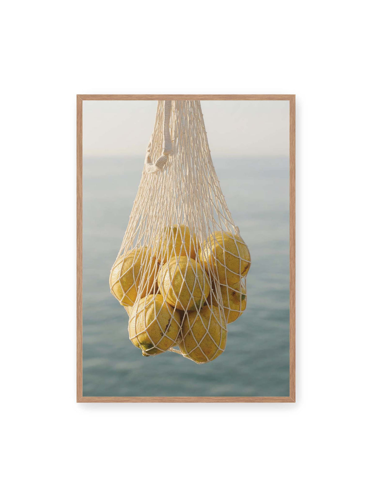 70x100 cm, Eichenrahmen – Gelbe Zitronen in einem hellen geknüpften Netz vor einer weich verschwimmenden blauen Wasserfläche.