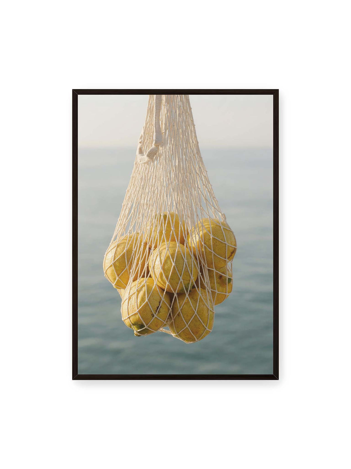 70x100 cm, schwarzer Rahmen – Gelbe Zitronen in einem hellen geknüpften Netz vor einer weich verschwimmenden blauen Wasserfläche.