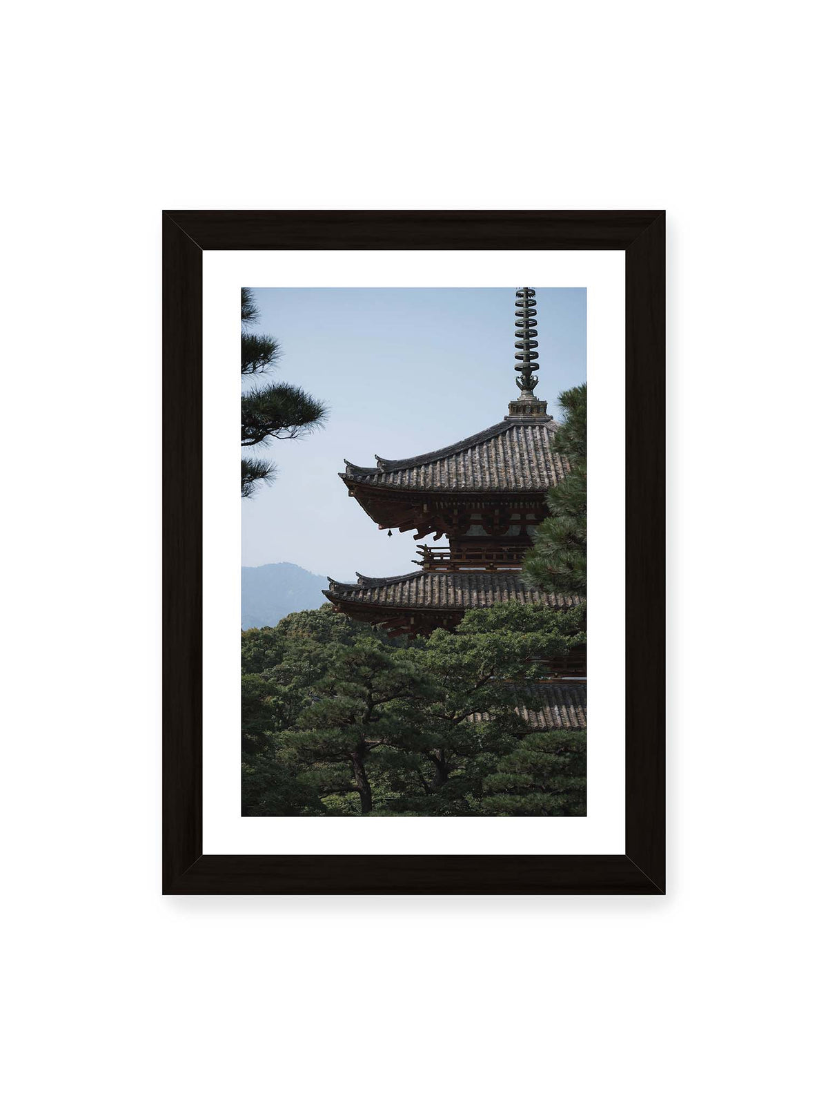 21x30 cm, weißer Galerie-Rand, schwarzer Rahmen – Mehrstufige Pagode mit geschwungenen Ziegeldächern zwischen dichten Kiefern vor einer blassen Bergsilhouette.