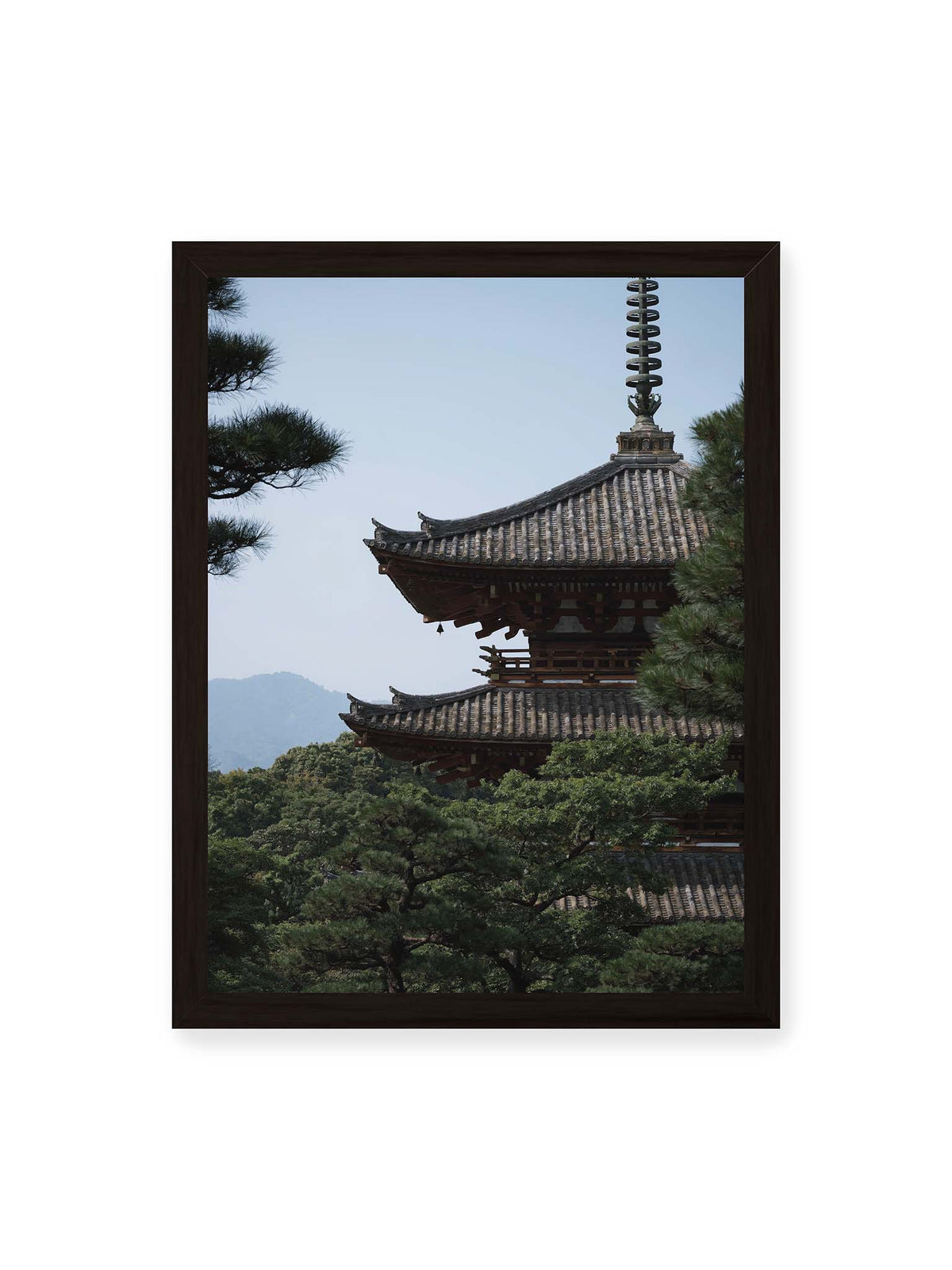 30x40 cm, schwarzer Rahmen – Mehrstufige Pagode mit geschwungenen Ziegeldächern zwischen dichten Kiefern vor einer blassen Bergsilhouette.