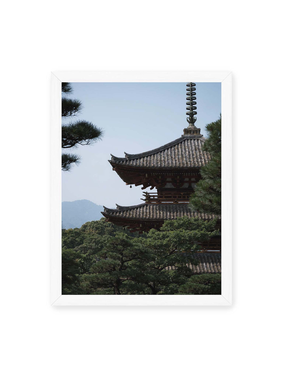 30x40 cm, weißer Rahmen – Mehrstufige Pagode mit geschwungenen Ziegeldächern zwischen dichten Kiefern vor einer blassen Bergsilhouette.