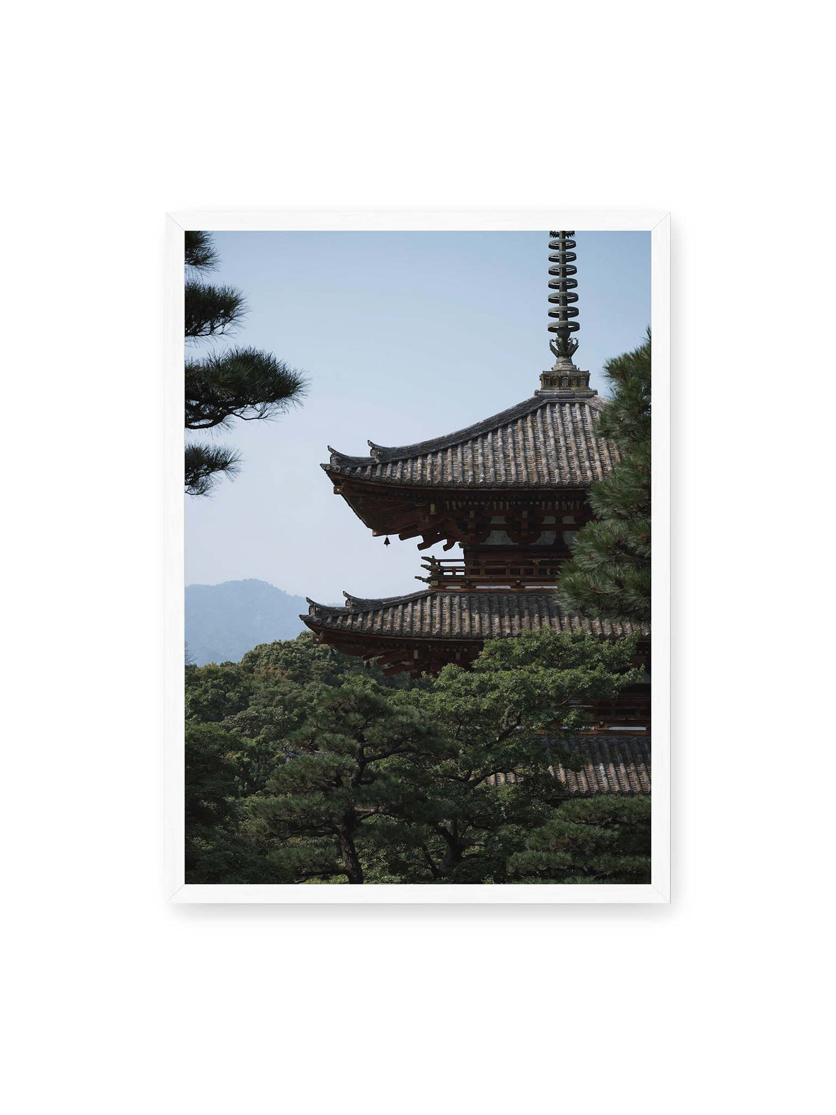 50x70 cm, weißer Rahmen – Mehrstufige Pagode mit geschwungenen Ziegeldächern zwischen dichten Kiefern vor einer blassen Bergsilhouette.