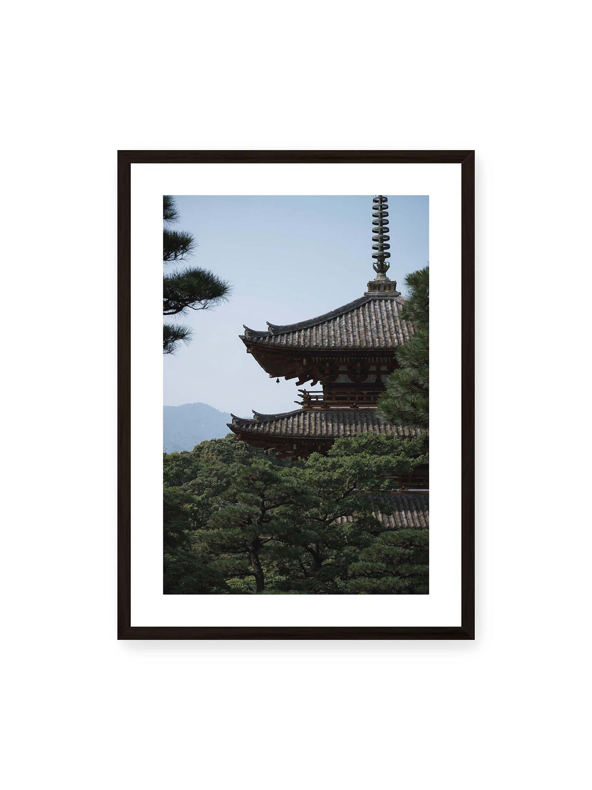 50x70 cm, weißer Galerie-Rand, schwarzer Rahmen – Mehrstufige Pagode mit geschwungenen Ziegeldächern zwischen dichten Kiefern vor einer blassen Bergsilhouette.