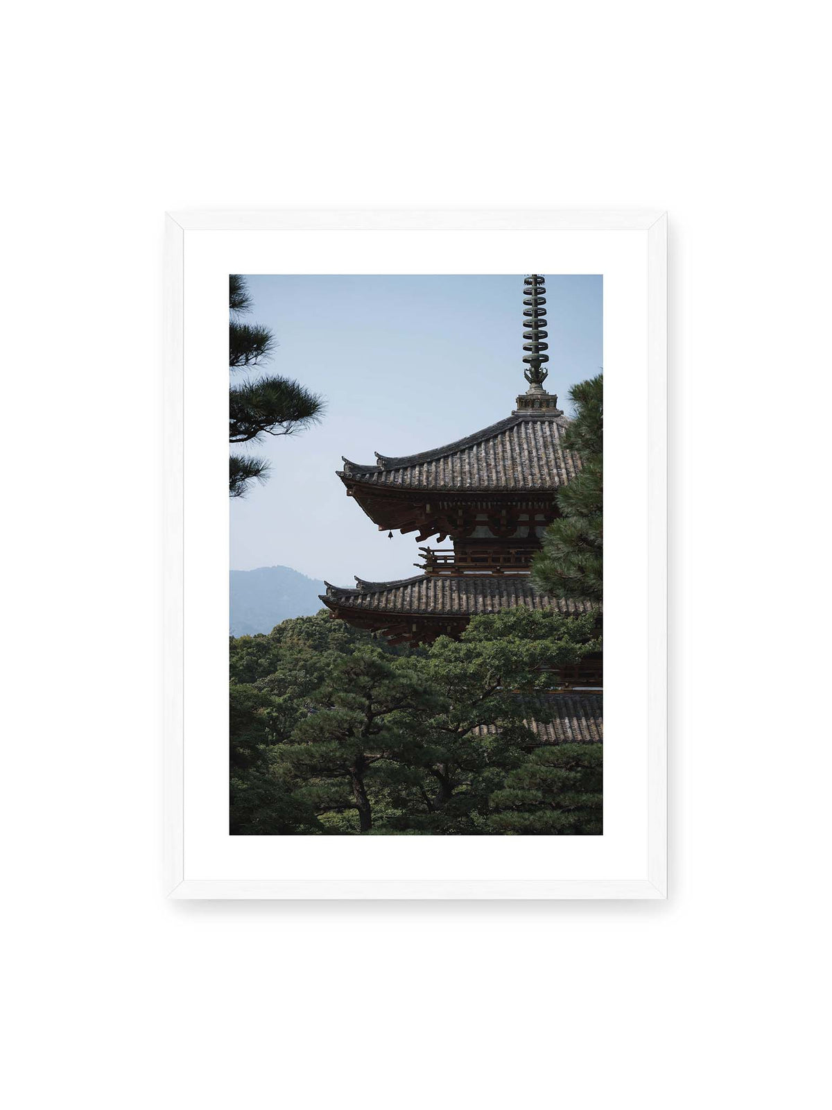 50x70 cm, weißer Galerie-Rand, weißer Rahmen – Mehrstufige Pagode mit geschwungenen Ziegeldächern zwischen dichten Kiefern vor einer blassen Bergsilhouette.