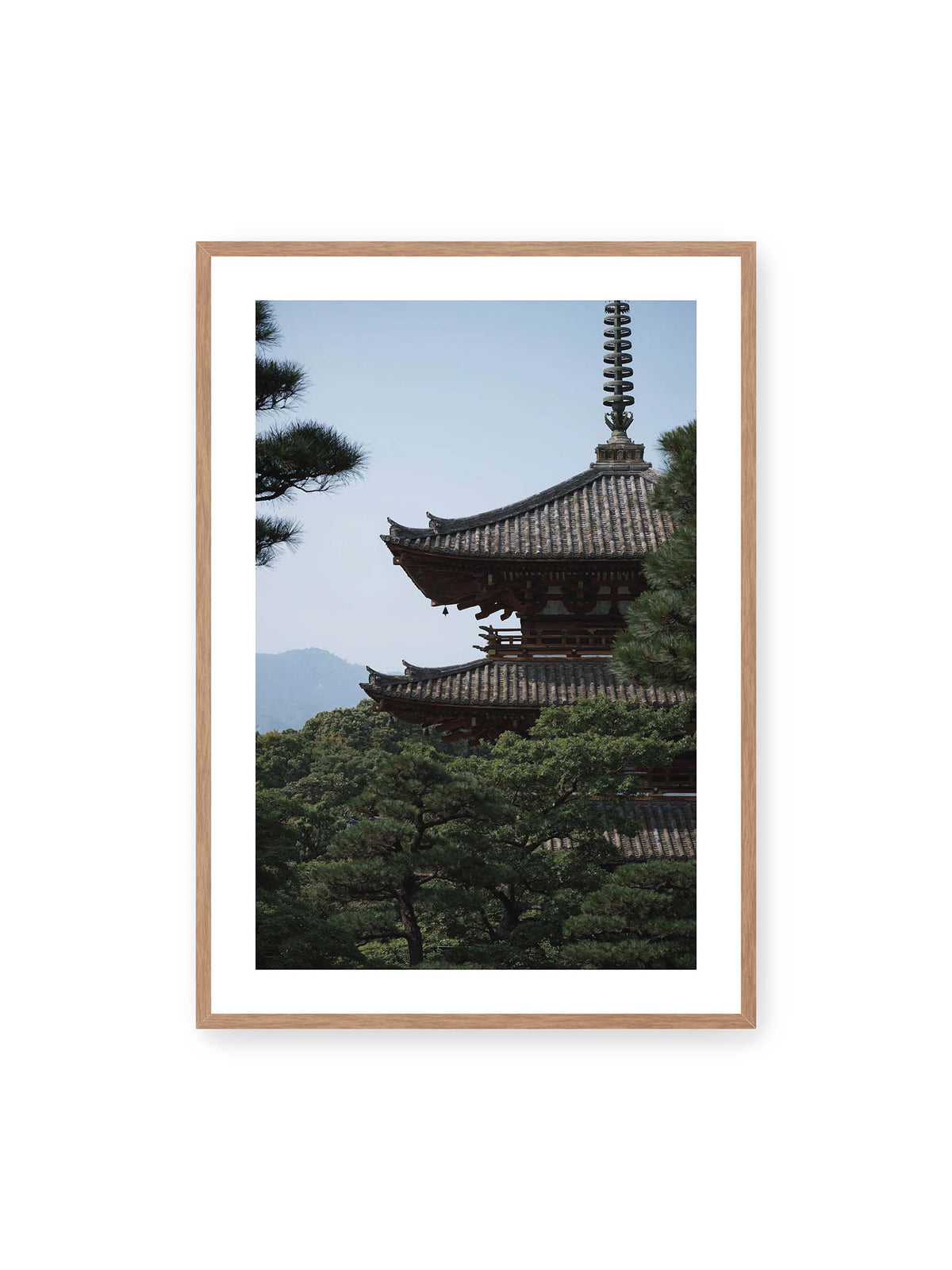 70x100 cm, weißer Galerie-Rand, Eichenrahmen – Mehrstufige Pagode mit geschwungenen Ziegeldächern zwischen dichten Kiefern vor einer blassen Bergsilhouette.
