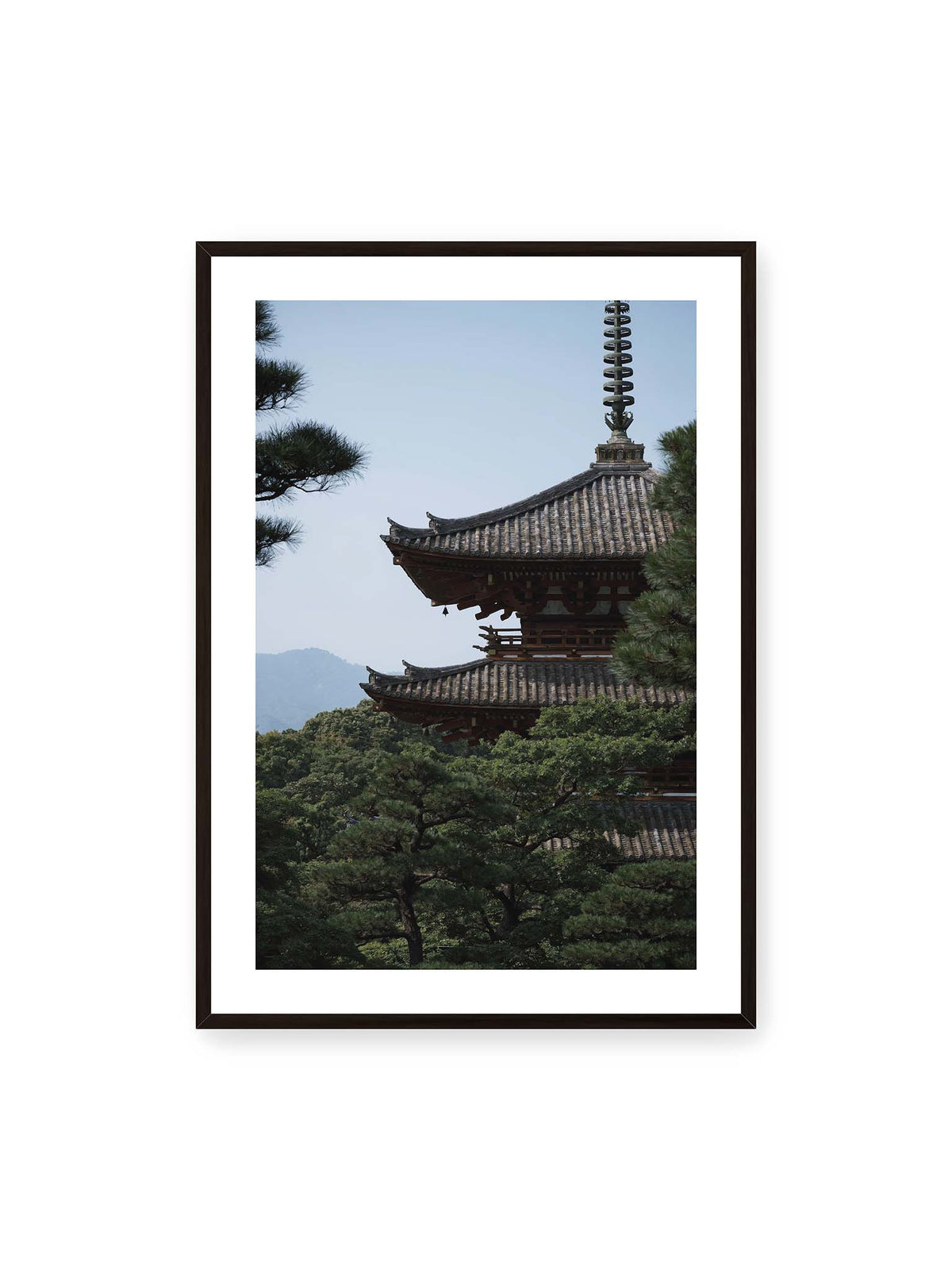 70x100 cm, weißer Galerie-Rand, schwarzer Rahmen – Mehrstufige Pagode mit geschwungenen Ziegeldächern zwischen dichten Kiefern vor einer blassen Bergsilhouette.