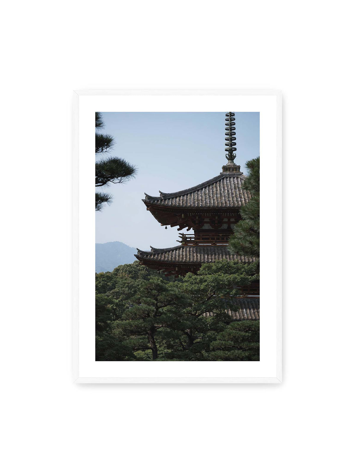 70x100 cm, weißer Galerie-Rand, weißer Rahmen – Mehrstufige Pagode mit geschwungenen Ziegeldächern zwischen dichten Kiefern vor einer blassen Bergsilhouette.
