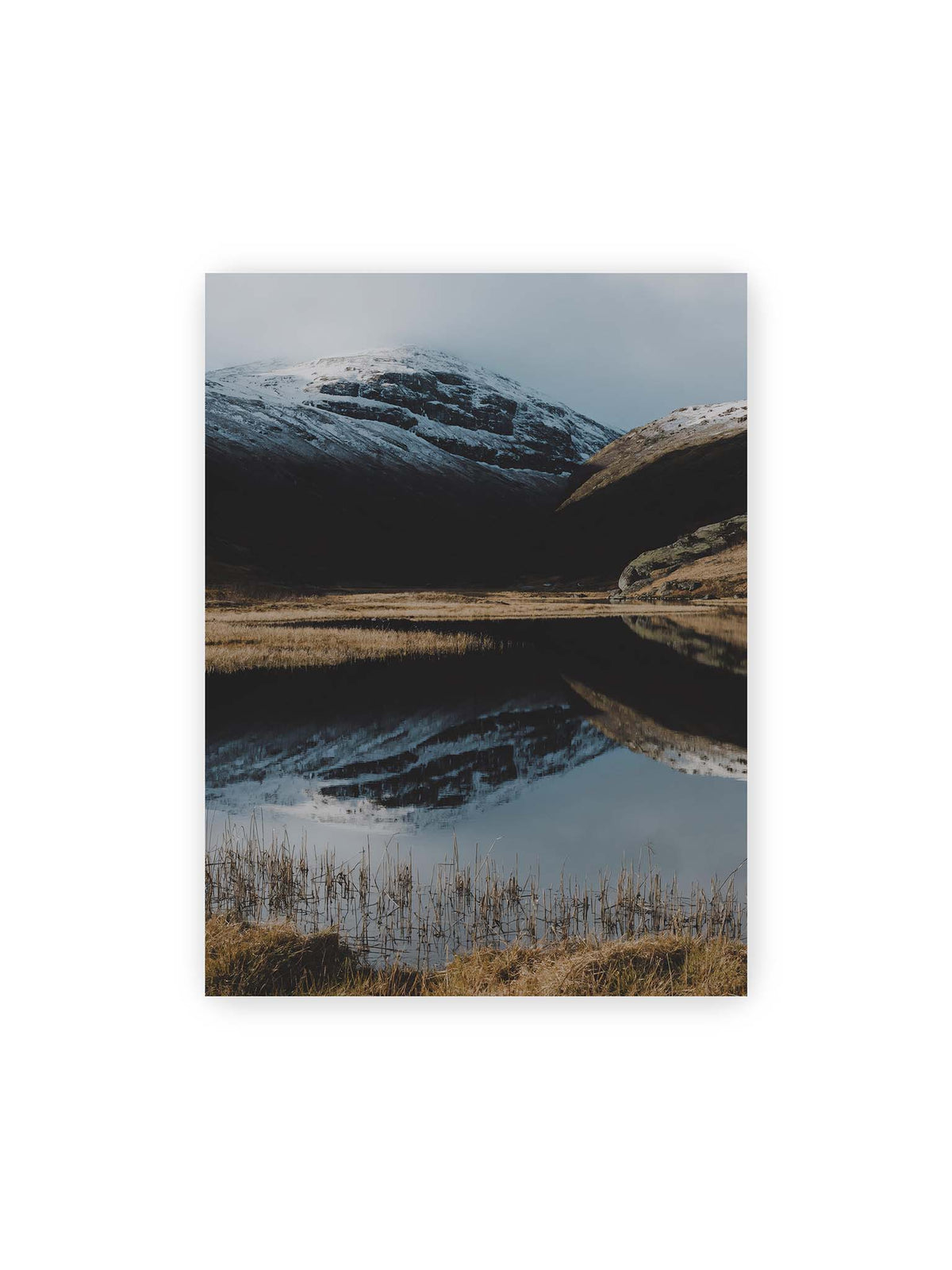 30x40 cm – Schneebedeckte Berghänge spiegeln sich in einem stillen Gewässer, umgeben von hellen Gräsern und Schilf.