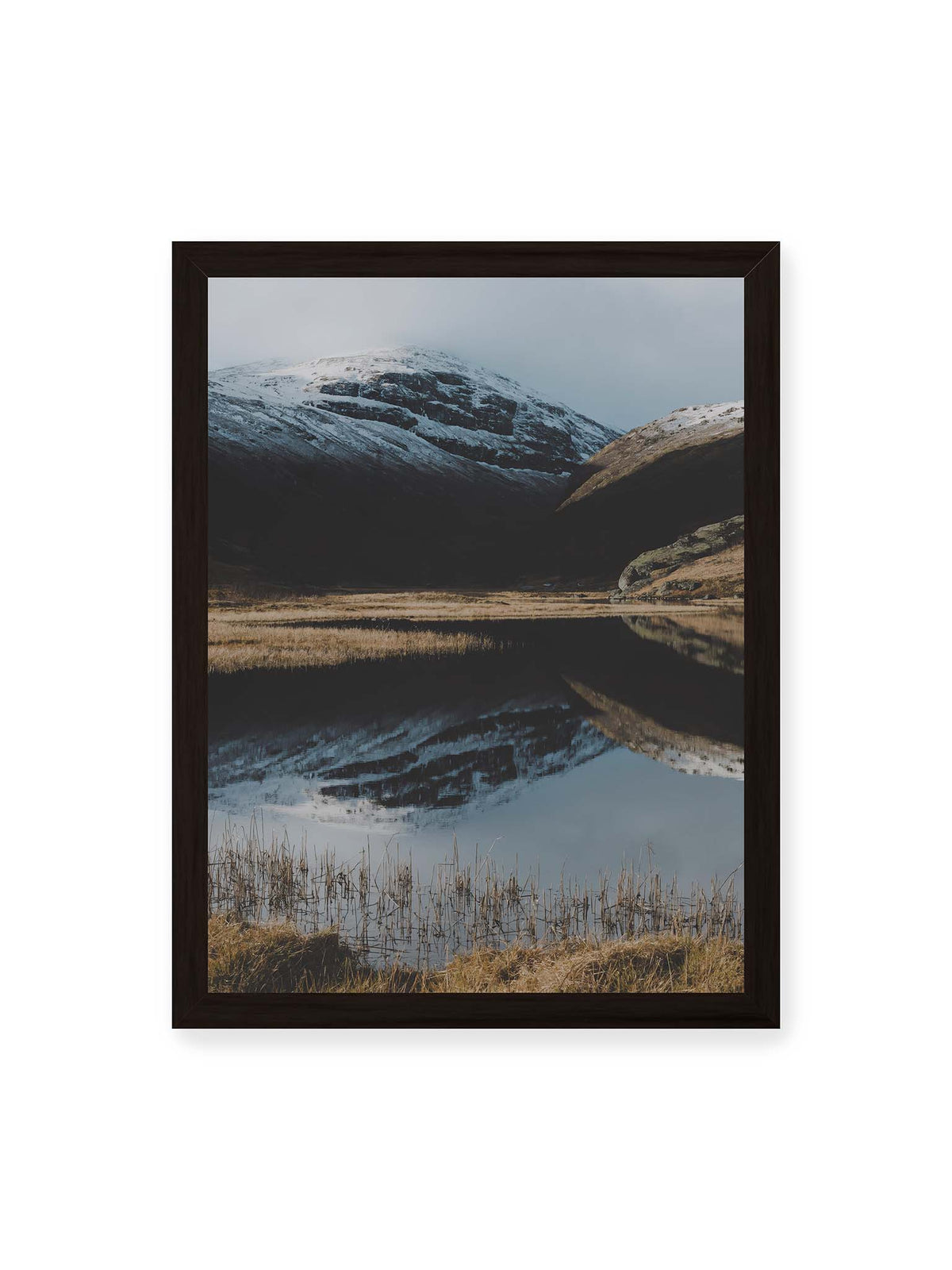 30x40 cm, schwarzer Rahmen – Schneebedeckte Berghänge spiegeln sich in einem stillen Gewässer, umgeben von hellen Gräsern und Schilf.