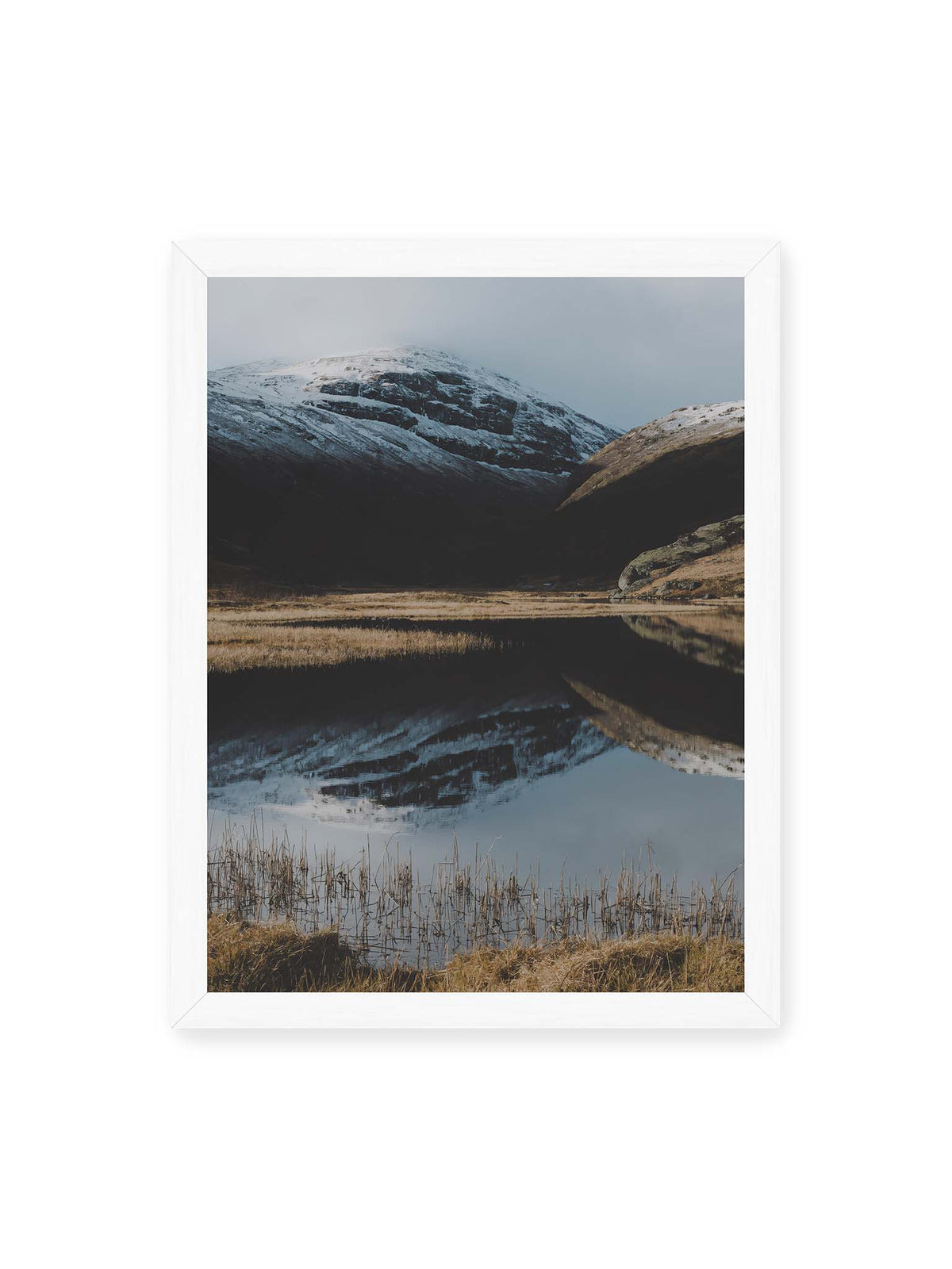 30x40 cm, weißer Rahmen – Schneebedeckte Berghänge spiegeln sich in einem stillen Gewässer, umgeben von hellen Gräsern und Schilf.