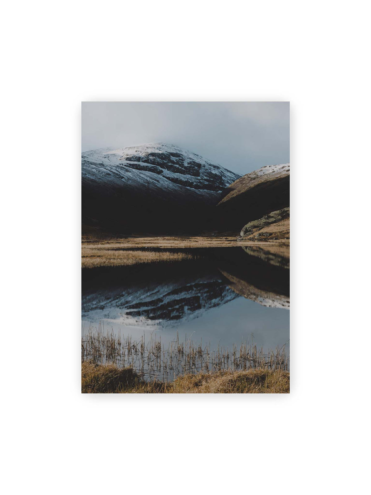 50x70 cm – Schneebedeckte Berghänge spiegeln sich in einem stillen Gewässer, umgeben von hellen Gräsern und Schilf.