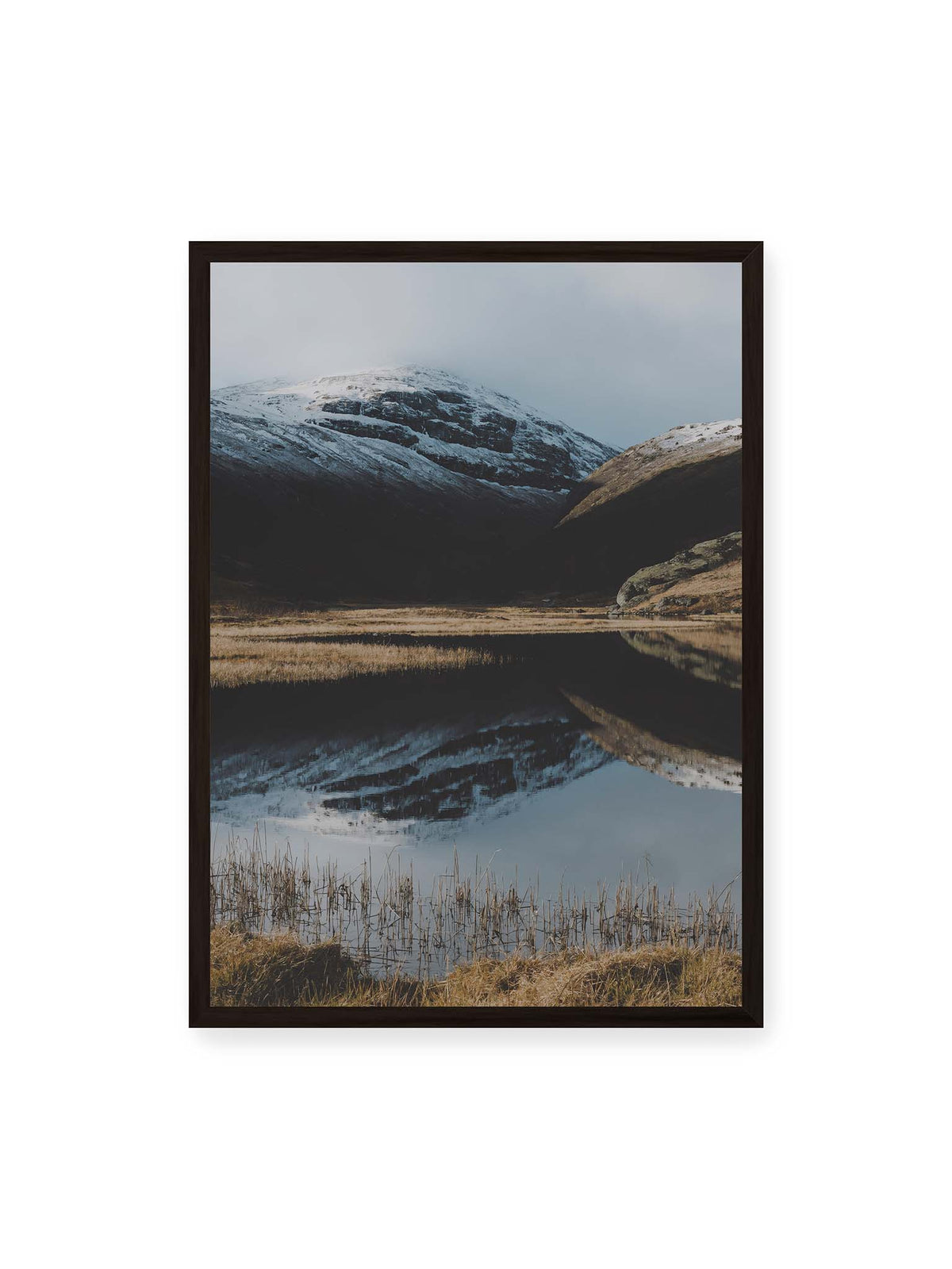 50x70 cm, schwarzer Rahmen – Schneebedeckte Berghänge spiegeln sich in einem stillen Gewässer, umgeben von hellen Gräsern und Schilf.