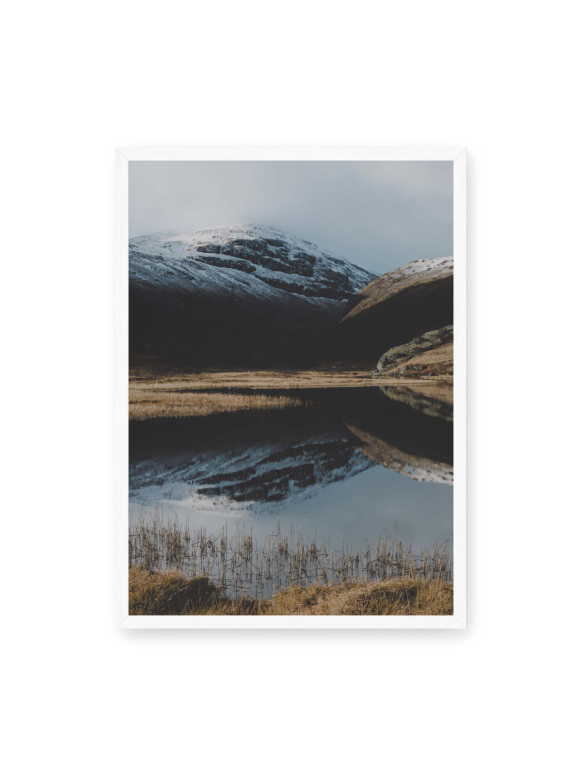 50x70 cm, weißer Rahmen – Schneebedeckte Berghänge spiegeln sich in einem stillen Gewässer, umgeben von hellen Gräsern und Schilf.