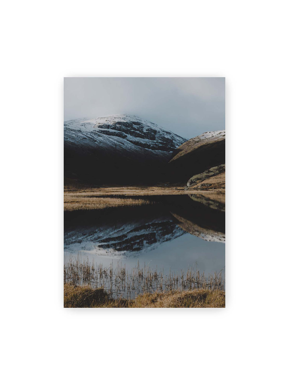 70x100 cm – Schneebedeckte Berghänge spiegeln sich in einem stillen Gewässer, umgeben von hellen Gräsern und Schilf.