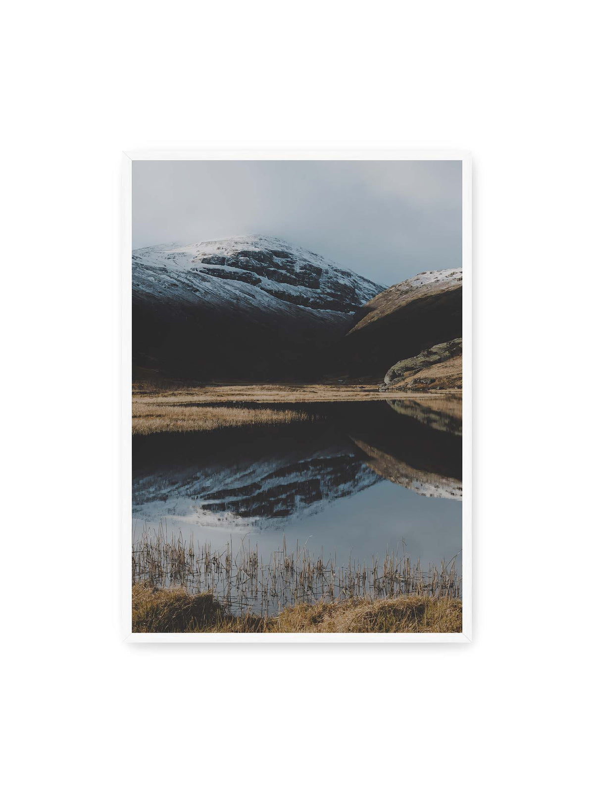 70x100 cm, weißer Rahmen – Schneebedeckte Berghänge spiegeln sich in einem stillen Gewässer, umgeben von hellen Gräsern und Schilf.