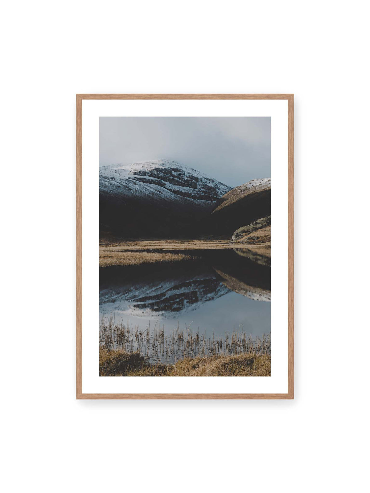 70x100 cm, weißer Galerie-Rand, Eichenrahmen – Schneebedeckte Berghänge spiegeln sich in einem stillen Gewässer, umgeben von hellen Gräsern und Schilf.