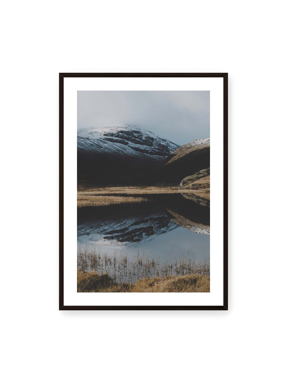 70x100 cm, weißer Galerie-Rand, schwarzer Rahmen – Schneebedeckte Berghänge spiegeln sich in einem stillen Gewässer, umgeben von hellen Gräsern und Schilf.