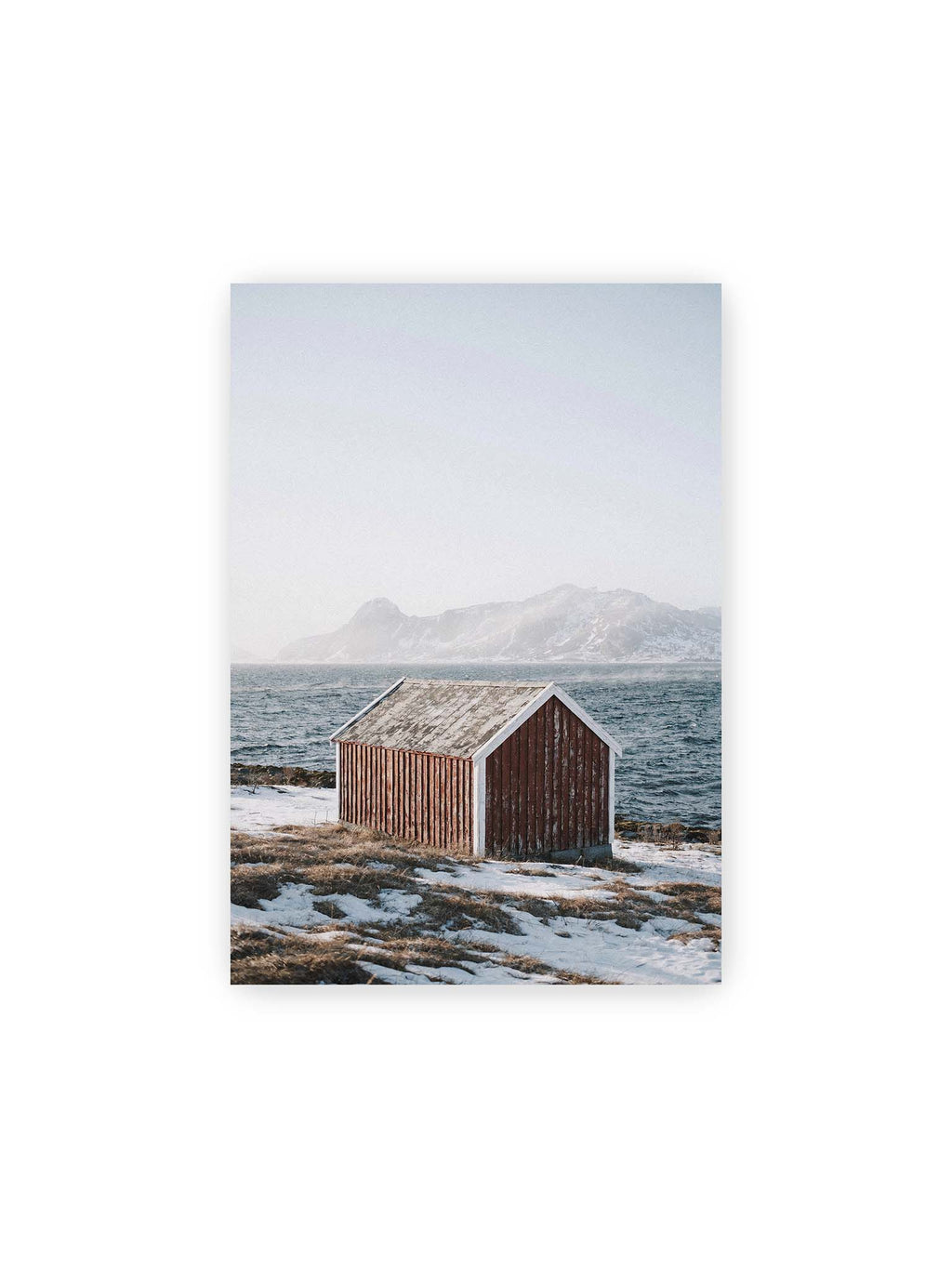 21x30 cm – Verwitterte rote Holzhütte an einer verschneiten Küste mit dunklem Wasser und felsigen Bergen im Hintergrund.