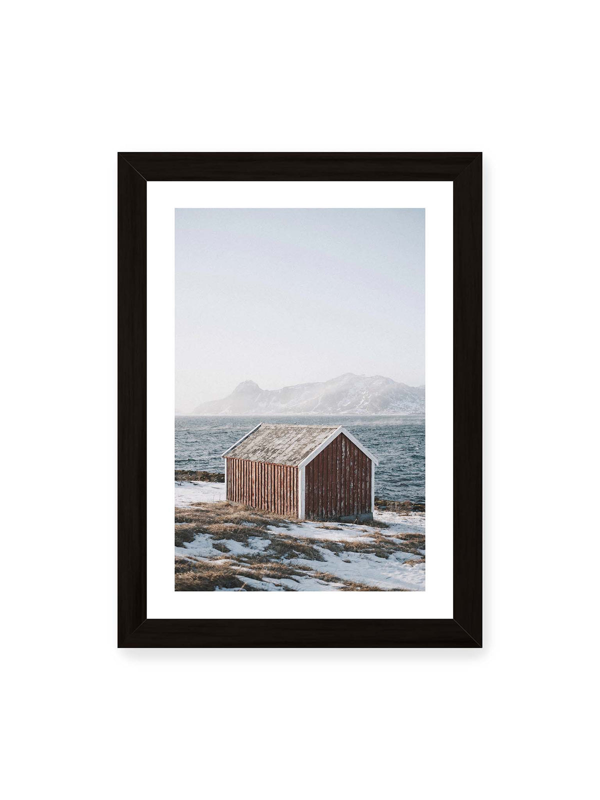 21x30 cm, weißer Galerie-Rand, schwarzer Rahmen – Verwitterte rote Holzhütte an einer verschneiten Küste mit dunklem Wasser und felsigen Bergen im Hintergrund.