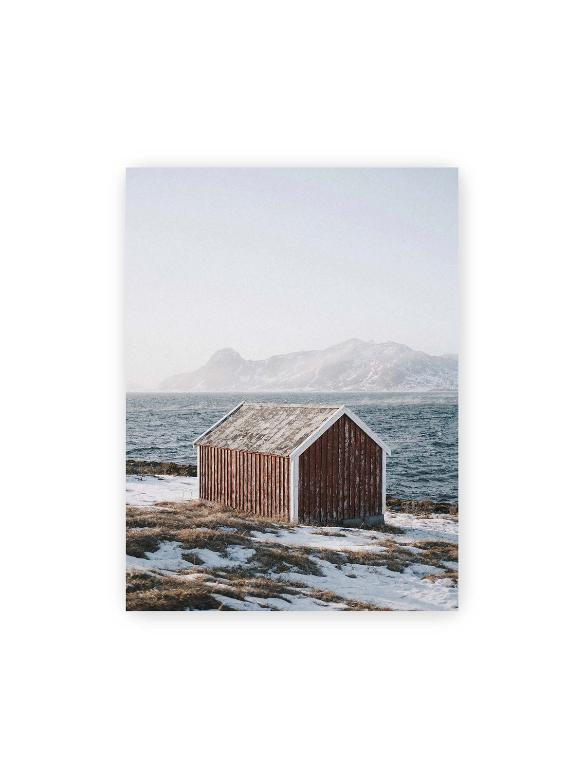 30x40 cm – Verwitterte rote Holzhütte an einer verschneiten Küste mit dunklem Wasser und felsigen Bergen im Hintergrund.