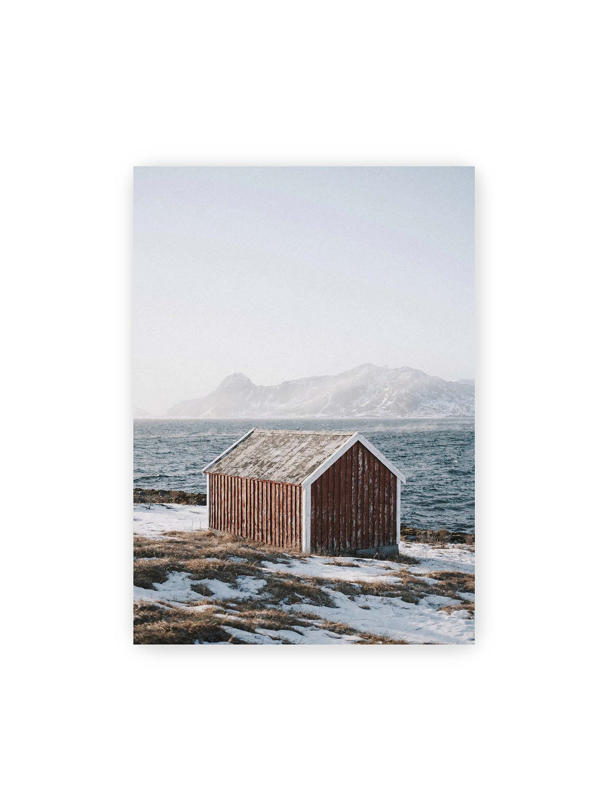50x70 cm – Verwitterte rote Holzhütte an einer verschneiten Küste mit dunklem Wasser und felsigen Bergen im Hintergrund.