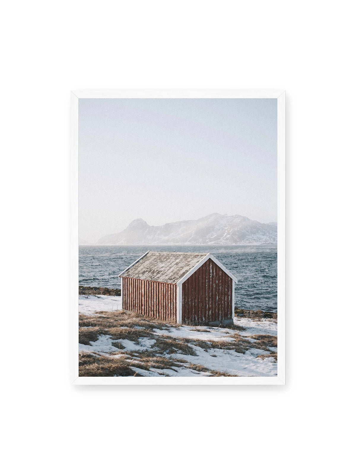 50x70 cm, weißer Rahmen – Verwitterte rote Holzhütte an einer verschneiten Küste mit dunklem Wasser und felsigen Bergen im Hintergrund.
