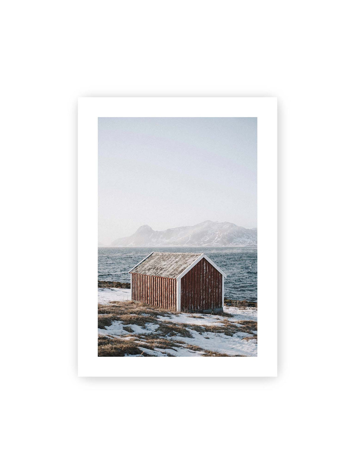 50x70 cm, weißer Galerie-Rand – Verwitterte rote Holzhütte an einer verschneiten Küste mit dunklem Wasser und felsigen Bergen im Hintergrund.