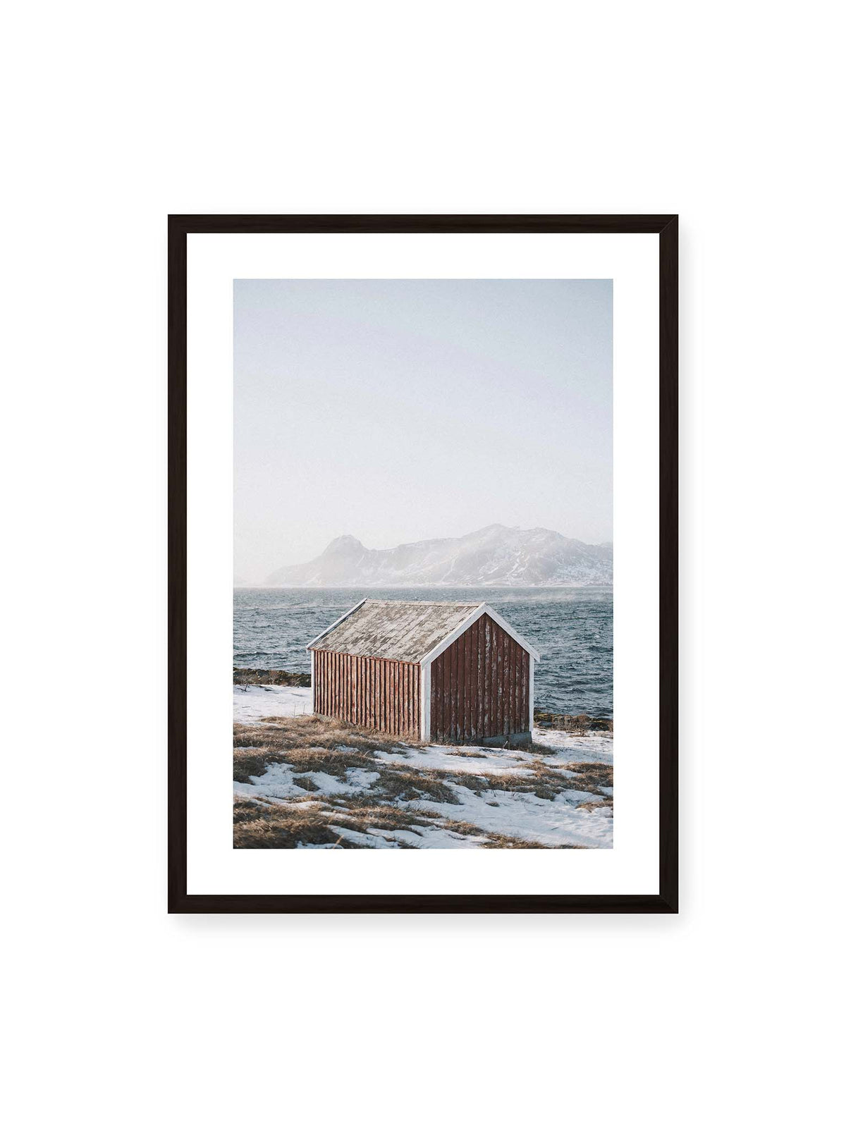 50x70 cm, weißer Galerie-Rand, schwarzer Rahmen – Verwitterte rote Holzhütte an einer verschneiten Küste mit dunklem Wasser und felsigen Bergen im Hintergrund.