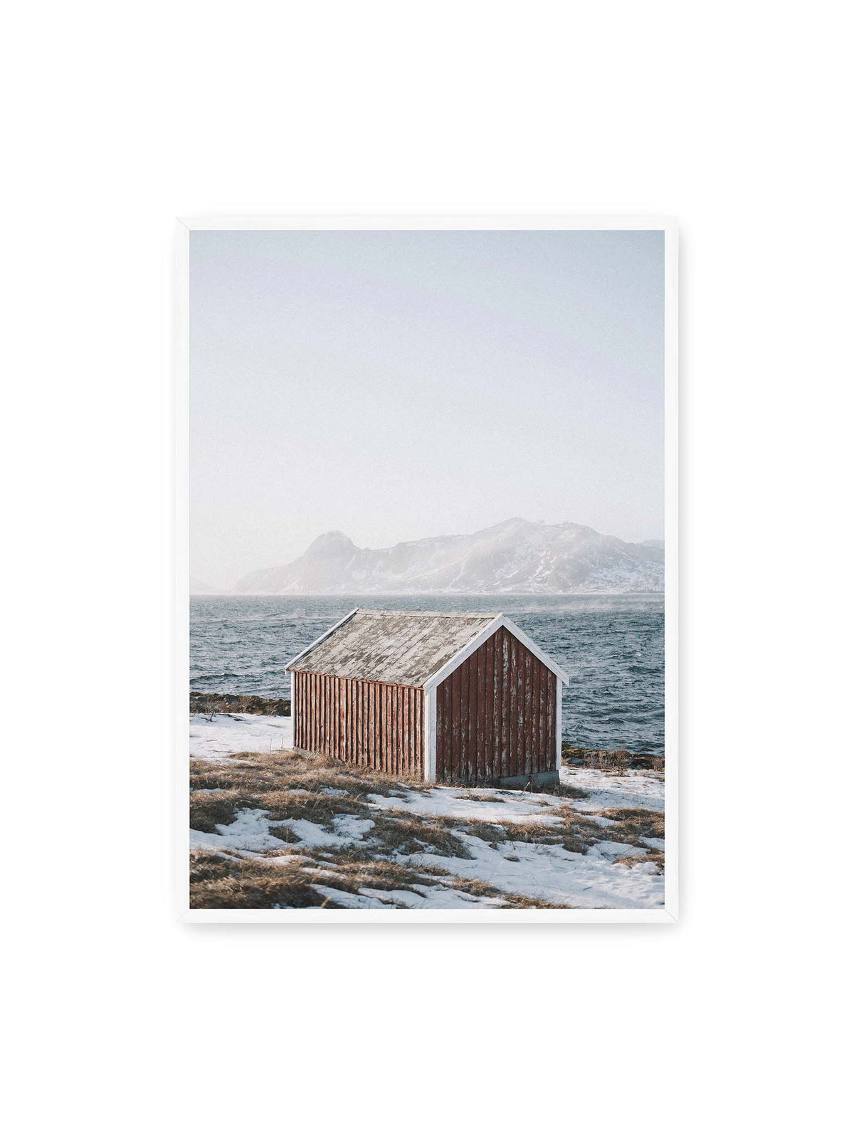 70x100 cm, weißer Rahmen – Verwitterte rote Holzhütte an einer verschneiten Küste mit dunklem Wasser und felsigen Bergen im Hintergrund.