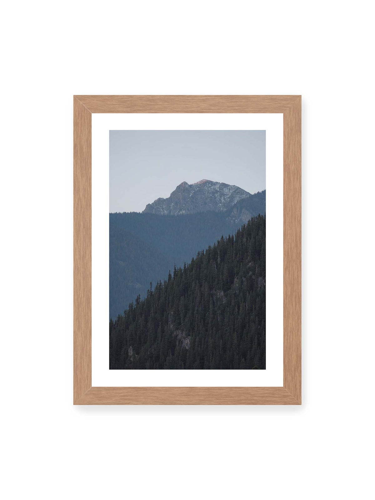21x30 cm, weißer Galerie-Rand, Eichenrahmen – Bewaldete Berghänge staffeln sich vor einem felsigen Gipfel mit einzelnen Schneeflächen und blaugrauem Himmel.