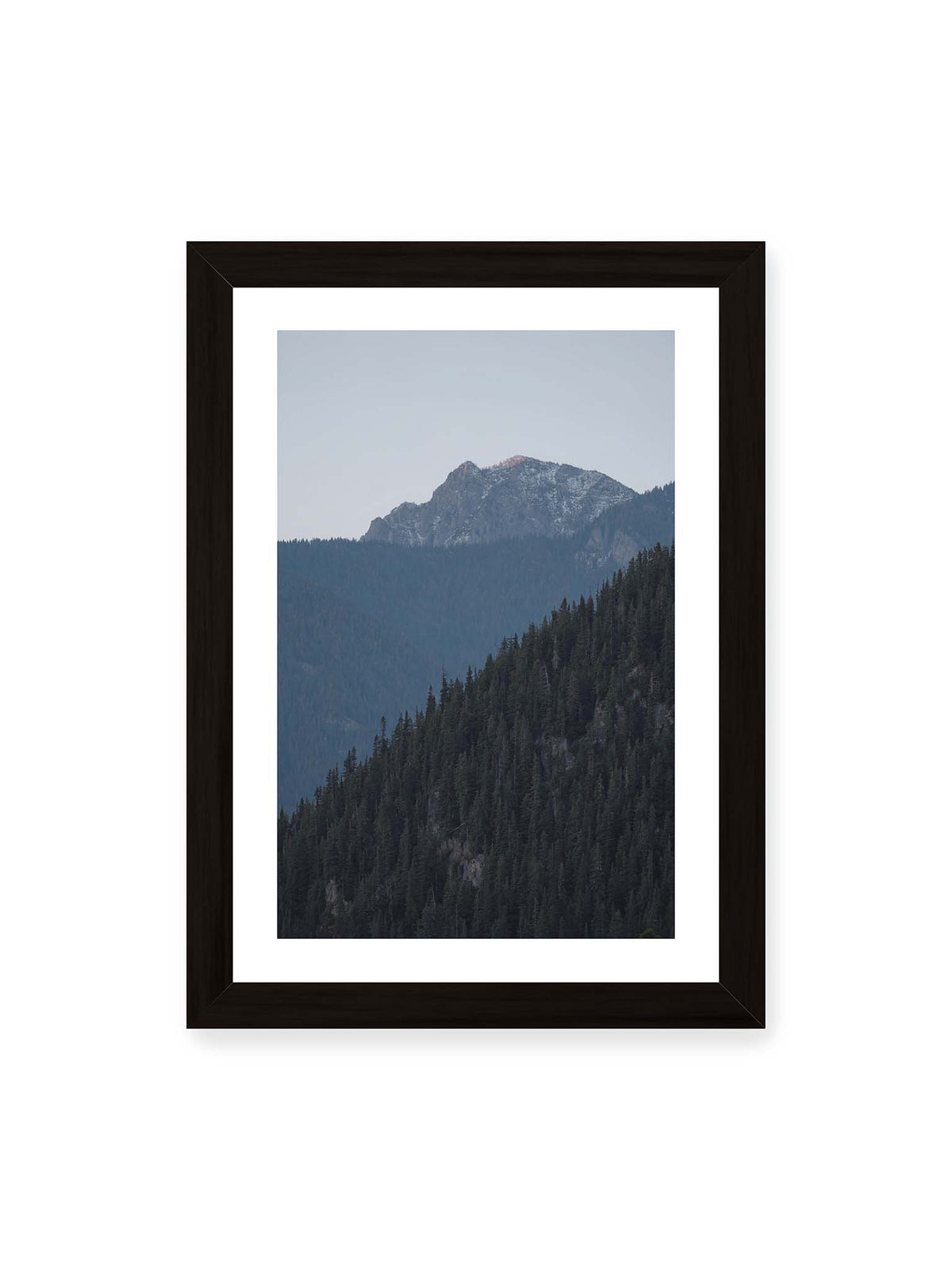 21x30 cm, weißer Galerie-Rand, schwarzer Rahmen – Bewaldete Berghänge staffeln sich vor einem felsigen Gipfel mit einzelnen Schneeflächen und blaugrauem Himmel.
