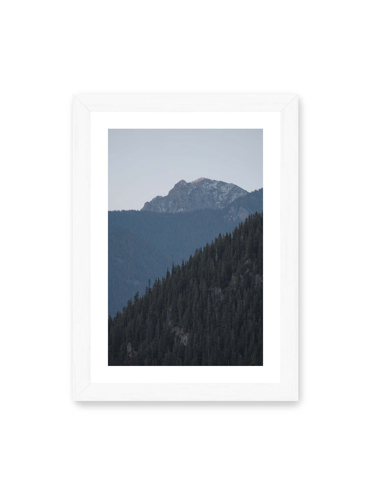 21x30 cm, weißer Galerie-Rand, weißer Rahmen – Bewaldete Berghänge staffeln sich vor einem felsigen Gipfel mit einzelnen Schneeflächen und blaugrauem Himmel.