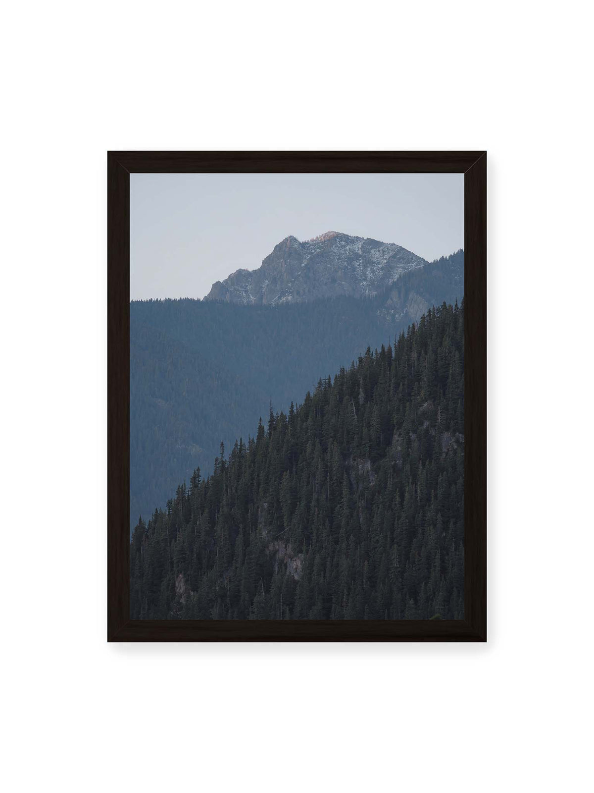30x40 cm, schwarzer Rahmen – Bewaldete Berghänge staffeln sich vor einem felsigen Gipfel mit einzelnen Schneeflächen und blaugrauem Himmel.