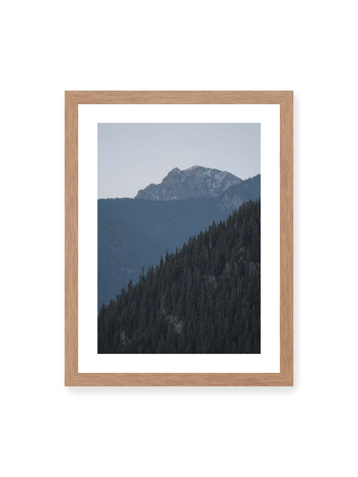 30x40 cm, weißer Galerie-Rand, Eichenrahmen – Bewaldete Berghänge staffeln sich vor einem felsigen Gipfel mit einzelnen Schneeflächen und blaugrauem Himmel.