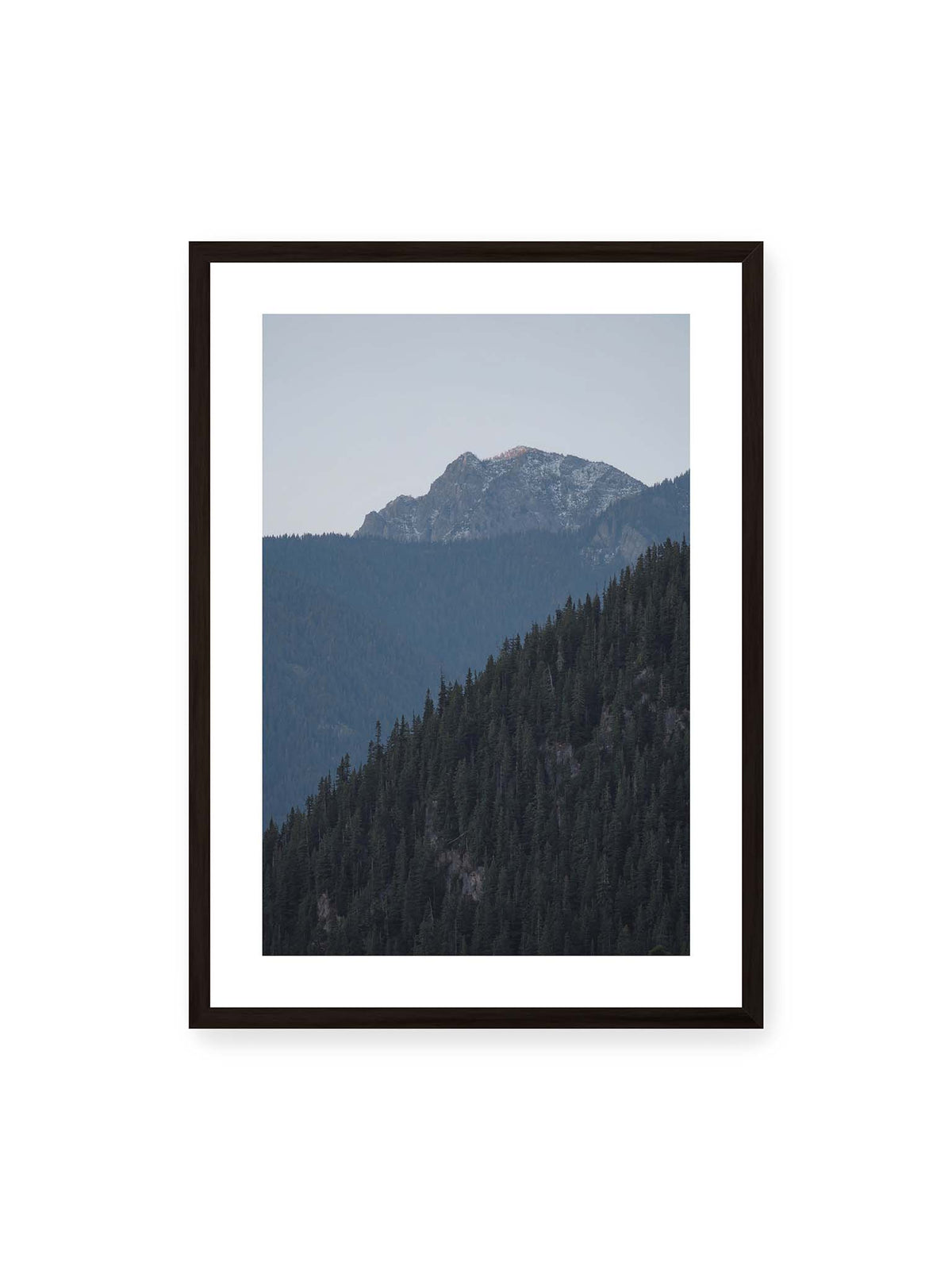 50x70 cm, weißer Galerie-Rand, schwarzer Rahmen – Bewaldete Berghänge staffeln sich vor einem felsigen Gipfel mit einzelnen Schneeflächen und blaugrauem Himmel.