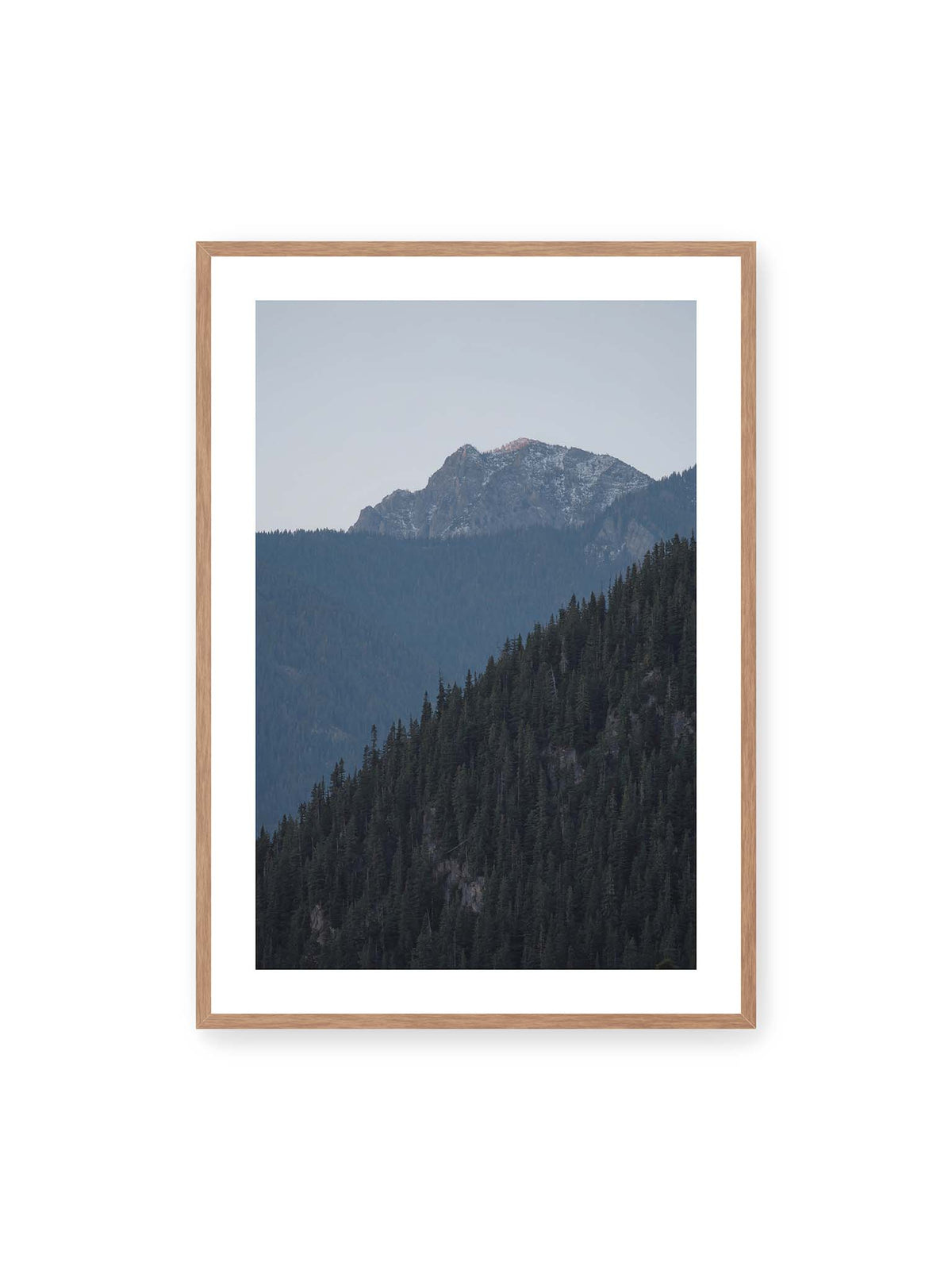 70x100 cm, weißer Galerie-Rand, Eichenrahmen – Bewaldete Berghänge staffeln sich vor einem felsigen Gipfel mit einzelnen Schneeflächen und blaugrauem Himmel.