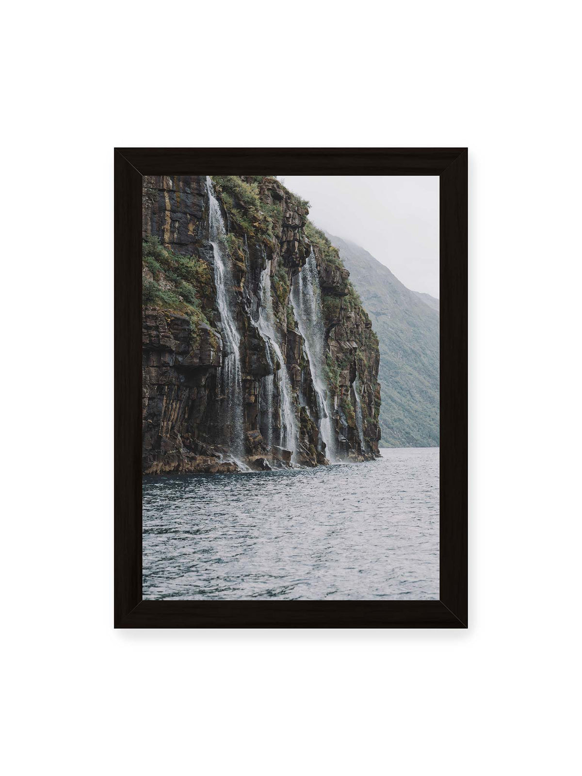 21x30 cm, schwarzer Rahmen – Mehrere Wasserfälle stürzen über eine dunkle, bewachsene Felswand direkt in eine weite Wasserfläche.