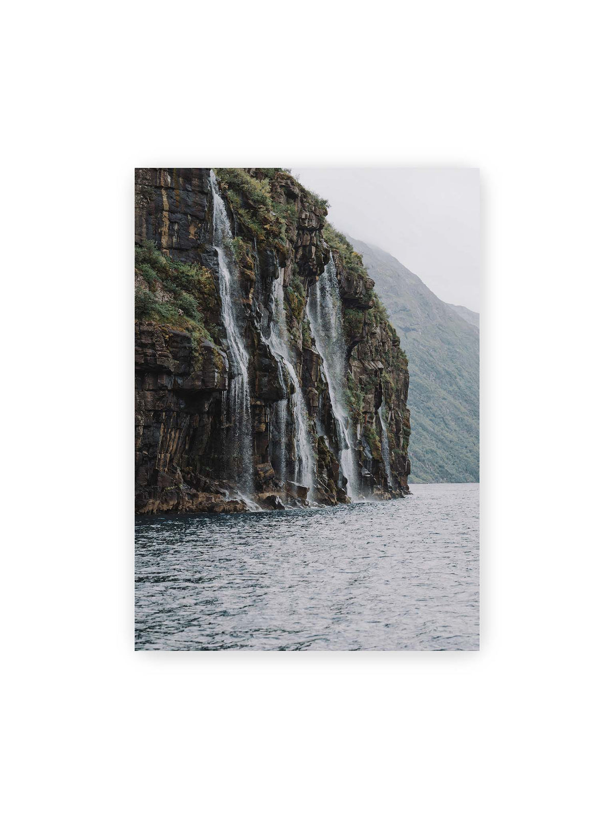 50x70 cm – Mehrere Wasserfälle stürzen über eine dunkle, bewachsene Felswand direkt in eine weite Wasserfläche.