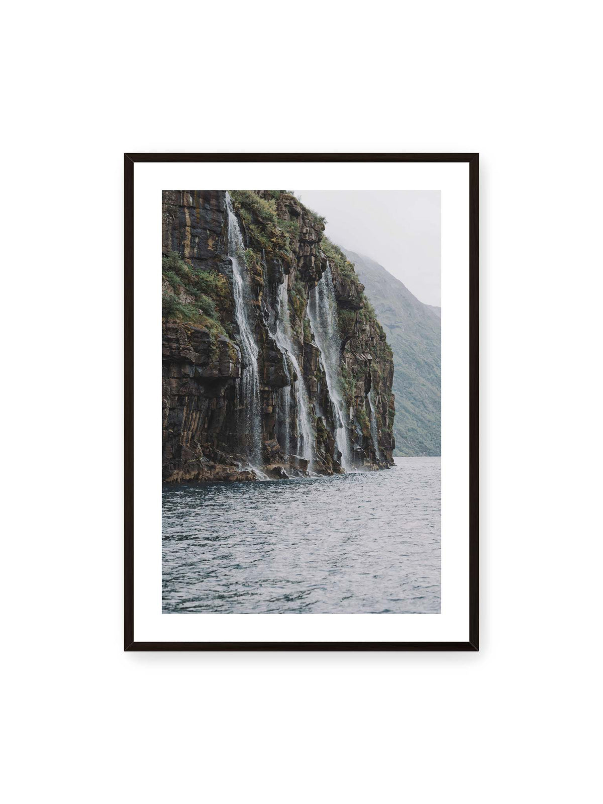 70x100 cm, weißer Galerie-Rand, schwarzer Rahmen – Mehrere Wasserfälle stürzen über eine dunkle, bewachsene Felswand direkt in eine weite Wasserfläche.