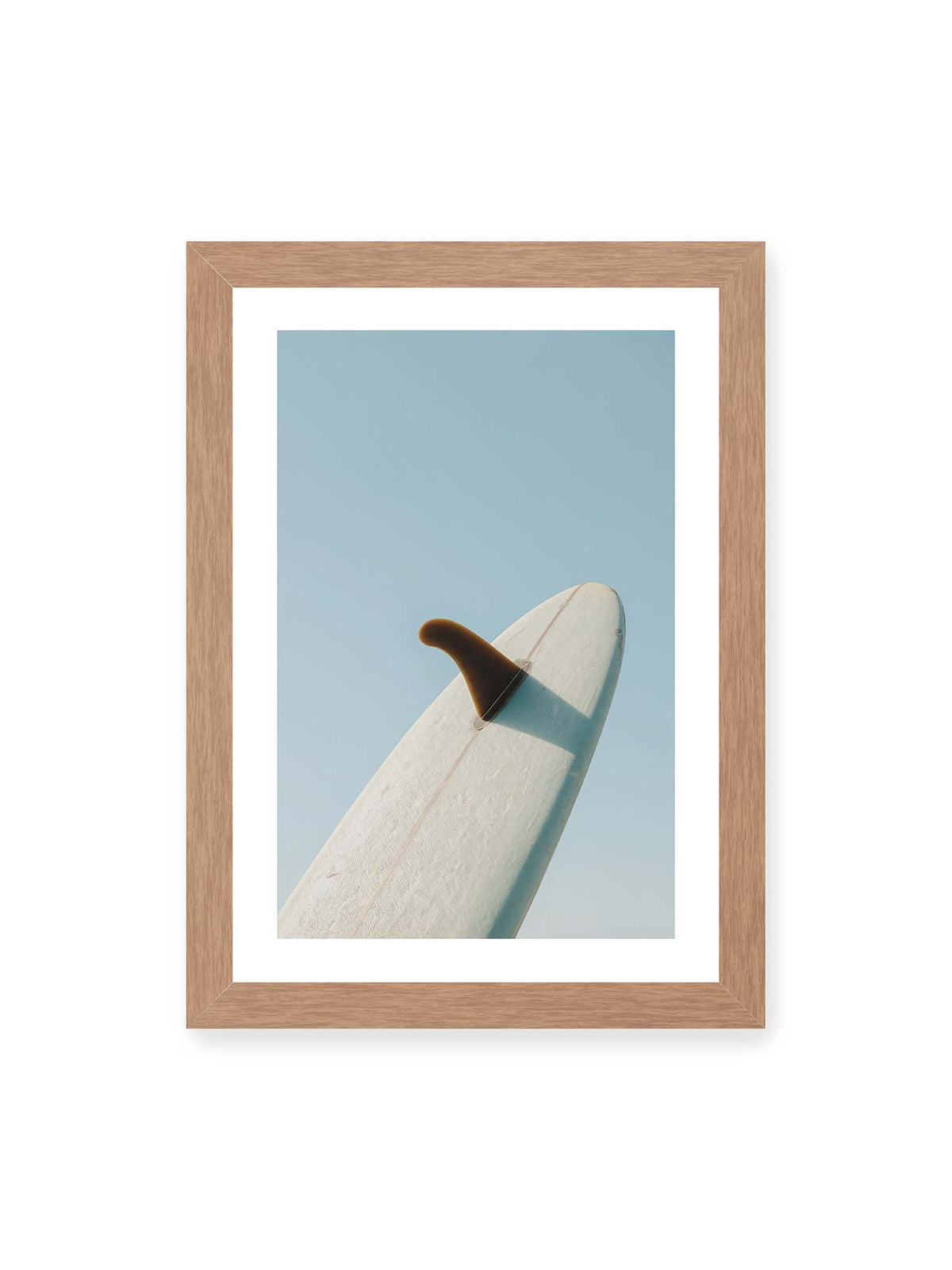 21x30 cm, weißer Galerie-Rand, Eichenrahmen – Helles Surfbrett mit dunkler Finne und markantem Schatten ragt diagonal vor einen klaren blauen Himmel.