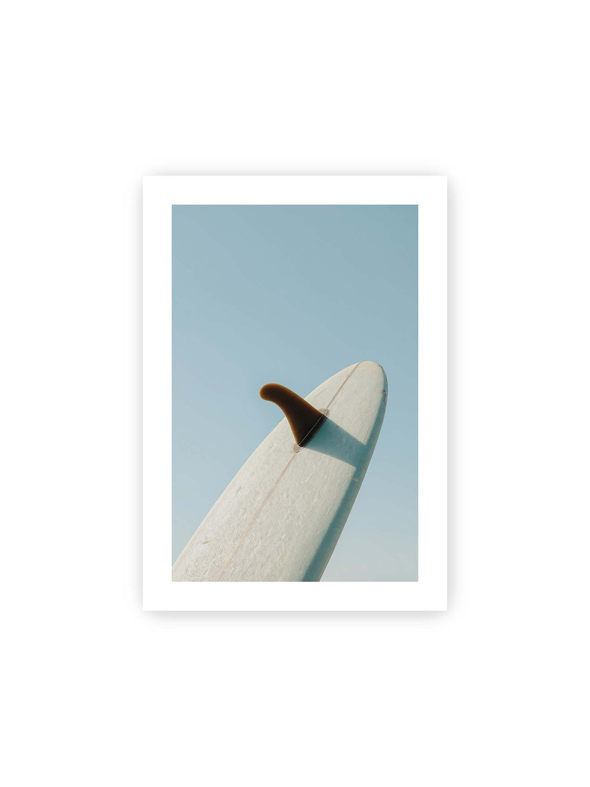 21x30 cm, weißer Galerie-Rand – Helles Surfbrett mit dunkler Finne und markantem Schatten ragt diagonal vor einen klaren blauen Himmel.