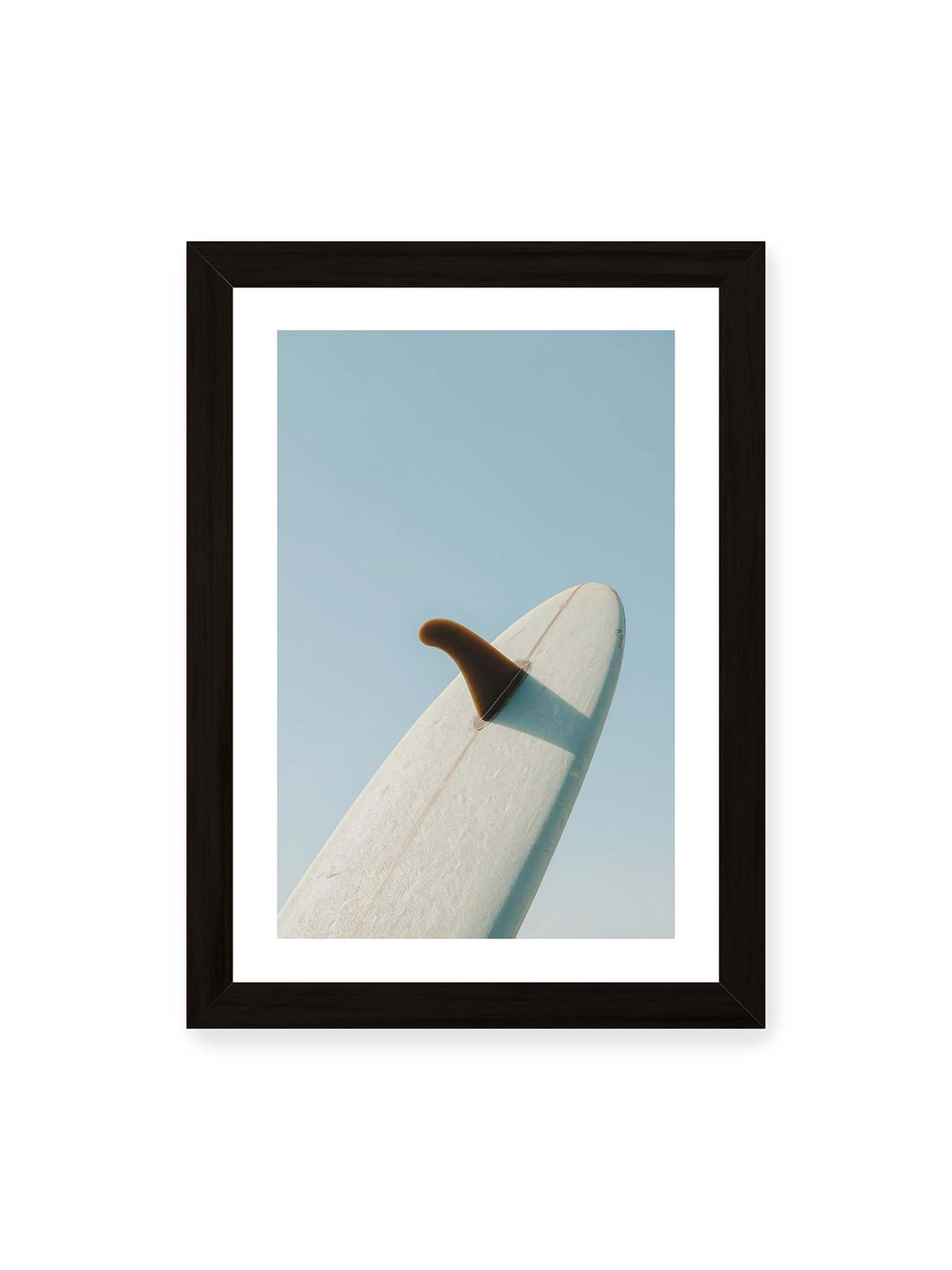 21x30 cm, weißer Galerie-Rand, schwarzer Rahmen – Helles Surfbrett mit dunkler Finne und markantem Schatten ragt diagonal vor einen klaren blauen Himmel.
