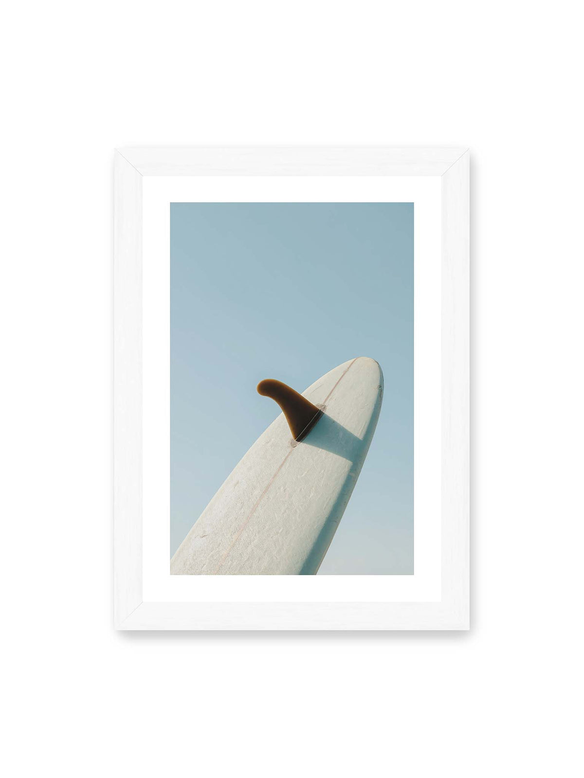 21x30 cm, weißer Galerie-Rand, weißer Rahmen – Helles Surfbrett mit dunkler Finne und markantem Schatten ragt diagonal vor einen klaren blauen Himmel.