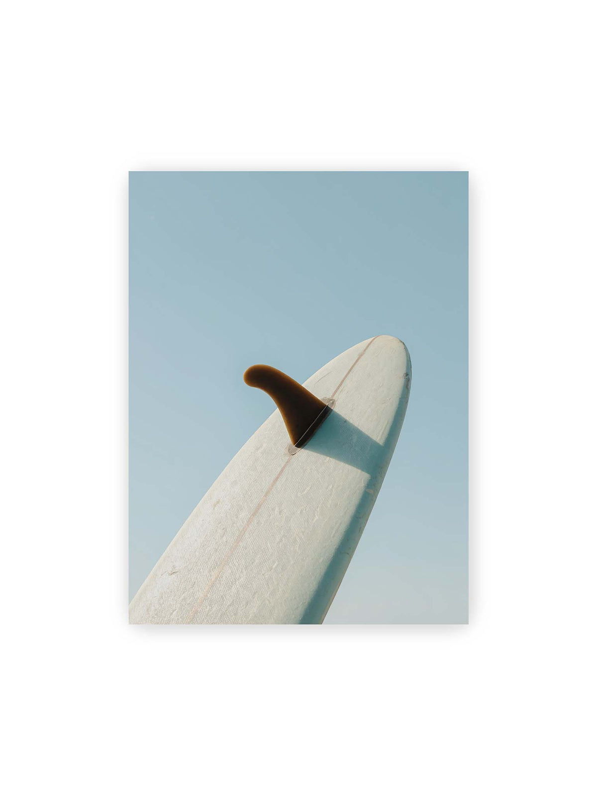 30x40 cm – Helles Surfbrett mit dunkler Finne und markantem Schatten ragt diagonal vor einen klaren blauen Himmel.