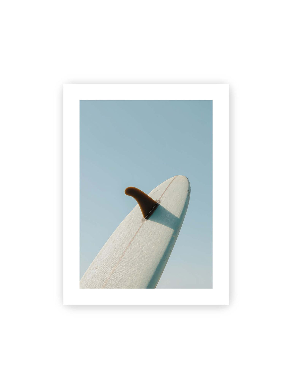 30x40 cm, weißer Galerie-Rand – Helles Surfbrett mit dunkler Finne und markantem Schatten ragt diagonal vor einen klaren blauen Himmel.